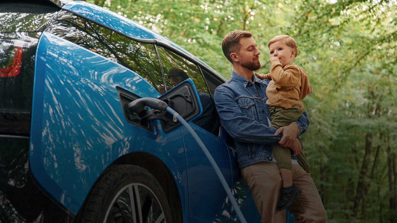 Ein Mann hat seinen Sohn am Arm. Sie stehen vor einem Elektroauto, das gerade geladen wird.
